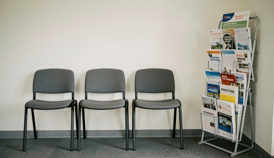 Illustrative image: a plain waiting area with two empty chairs, an information leaflet rack half full against a pale wall