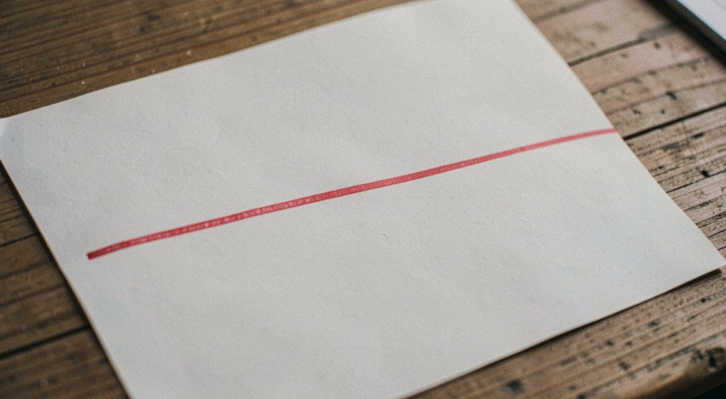 Illustrative image: a single ruled line drawn in red ink across a sheet of otherwise blank writing paper on a wooden surface